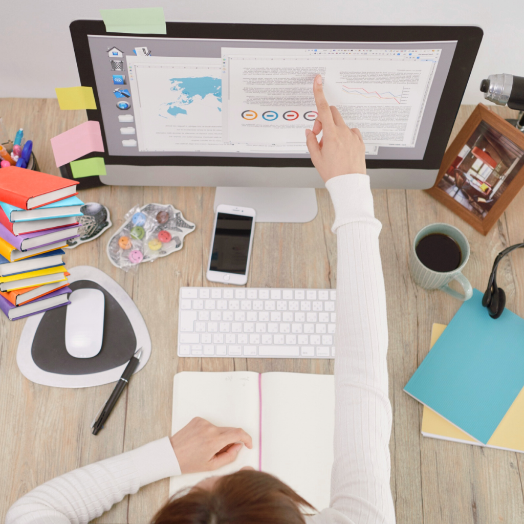 Eine Person zeigt etwas auf einem Computer, im Hintergrund steht ein Bürotisch mit mehreren Brüchern.